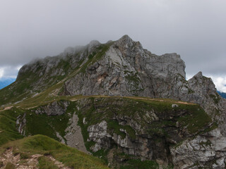 A panoramic alpine landscape of Montgart in the Alps featuring rugged rocky cliffs, soft green slopes, a winding mountain road and a dramatic cloudy sky