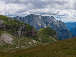 Fototapeta premium A panoramic alpine landscape of Montgart in the Alps featuring rugged rocky cliffs, soft green slopes, a winding mountain road and a dramatic cloudy sky