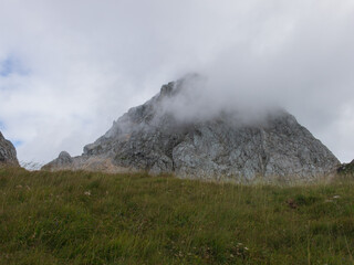 A panoramic alpine landscape of Montgart in the Alps featuring rugged rocky cliffs, soft green slopes, a winding mountain road and a dramatic cloudy sky