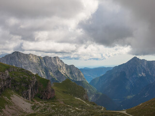 A panoramic alpine landscape of Montgart in the Alps featuring rugged rocky cliffs, soft green slopes, a winding mountain road and a dramatic cloudy sky