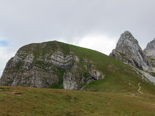 A panoramic alpine landscape of Montgart in the Alps featuring rugged rocky cliffs, soft green slopes, a winding mountain road and a dramatic cloudy sky