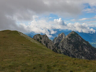 A panoramic alpine landscape of Montgart in the Alps featuring rugged rocky cliffs, soft green slopes, a winding mountain road and a dramatic cloudy sky