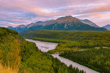 Fototapeta premium A winding glacial river cuts through dense boreal forest at golden hour along the Glenn Highway from Valdez to Anchorage, with sunset-lit Chugach Mountains glowing under pastel skies in early fall.