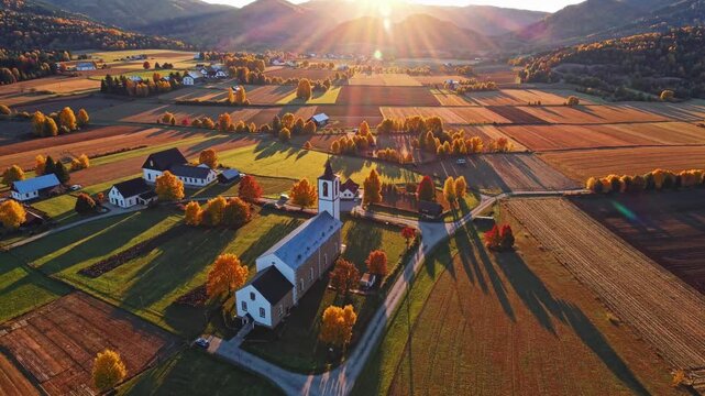 A quaint village is surrounded by colorful autumn foliage, with a church standing proudly amidst the vibrant trees. The setting sun casts long shadows over the rolling fields, enha