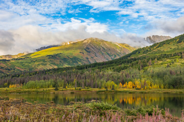 Obraz premium A serene lake reflects green mountains and soft clouds along Highway AK-1 South toward Seward, Alaska. The vibrant vegetation and peaceful water create a tranquil late-summer and early-fall mountain l