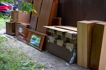 Dumped old outdoor furniture. Wooden cabinets, wooden doors, mirrors and shelves stacked against the wall of a city dumpster