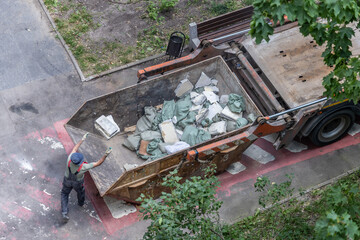 Overhead view of male worker managing construction waste disposal, caucasian, mature, in a large metallic dumpster filled with debris bags and building materials