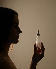 Closeup silhouette of a woman's profile holding a frosted glass serum dropper bottle with soft rim lighting on a beige studio background