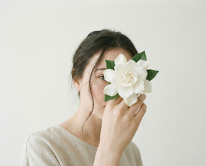 Woman's face partially hidden by a single white gardenia flower held close to her eye, minimalist natural beauty portrait on a bright background