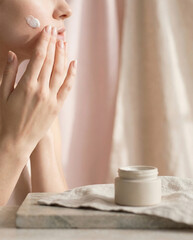 Minimal luxury beauty shot of a woman's hands applying white cream to her cheek next to a jar on a stone slab with linen and sheer curtains