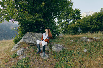 Naklejka premium Woman sitting on rock in mountain meadow under trees, looking up relaxed. Outdoor portrait with boots and coat, natural landscape, greenery and rocky foreground and sunlight.