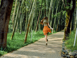 Woman running along a gravel path through a dense bamboo forest wearing a straw hat and orange skirt, enjoying summer nature trail, motion and peaceful green scenery.