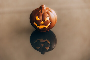 A small smiling pumpkin with a carved face is artistically lit from within against a reflective...