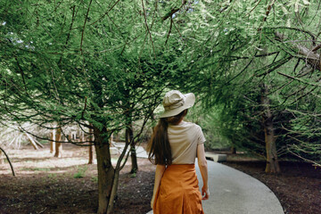 Naklejka premium Woman walking along a curved path in a leafy park under tall trees, wearing a sunhat and orange skirt. Nature scene in summer with peaceful, leisurely outdoor mood and soft sunlight.