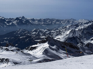Winter panoramic view of the snowy Alpine mountain range with dramatic peaks and deep valleys under a clear blue sky. Scenic high-altitude landscape ideal for travel and nature themes.