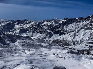 Winter panoramic view of the snowy Alpine mountain range with dramatic peaks and deep valleys under a clear blue sky. Scenic high-altitude landscape ideal for travel and nature themes.