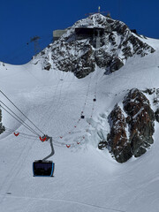 Cable car moving above a snowy mountain ridge in the Swiss Alps on a bright winter day. High-altitude alpine landscape with rugged peaks, blue sky and winter resort atmosphere.