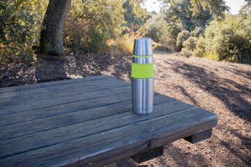 A stainless steel thermos with green fluor rubber rests on a rustic wooden picnic table in a sunny forest. It symbolizes hydration, sustainability, travel, hiking, and enjoying a healthy break on a