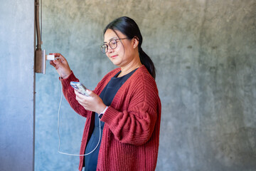 A woman plugging a charger into a wall outlet while holding her smartphone, illustrating modern technology use, connectivity, and daily digital habits in a simple indoor environment.