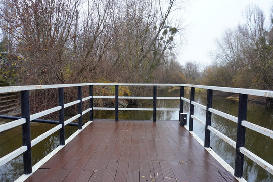 A wooden viewing platform with white and black railings extends over a calm river surrounded by bare trees under an overcast sky.