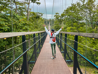 Rear view of a single female walking across a Suspension Bridge High in the Sky. She has her arms raised in the air. A forest of green trees surrounds her. Located in Northern Michigan, USA.