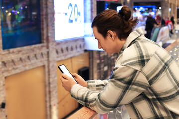 Young adult leaning on a railing in a shopping mall, looking intently at their smartphone; sale sign in background.