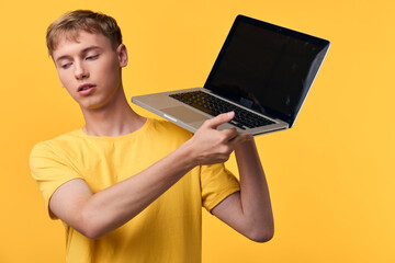 Male in yellow t shirt holding a laptop against a vibrant yellow background, emphasizing portable technology, digital work, and casual online learning in a modern studio lifestyle setup
