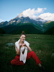 Naklejka premium Woman in red pants sits in a meadow with mountains behind, smiling and relaxed, enjoying outdoor nature, casual pose, scenic landscape for travel, wellness and hiking