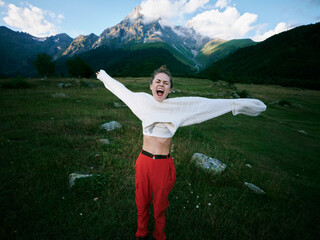 Naklejka premium Outdoor celebration in alpine landscape, a joyful figure spans arms wide, wearing a white sweater and red pants, with mountains and green valley in the background