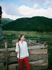 Naklejka premium Woman, countryside, outdoor, rural, standing beside a weathered fence with a staff, white sweater and red pants, expansive meadow and distant mountains under a bright sky