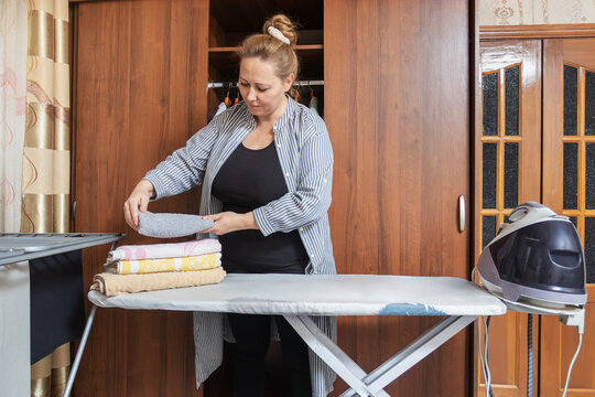 Mid-adult woman standing and ironing a white shirt using a modern steam generator iron on a board in a domestic laundry room setting.