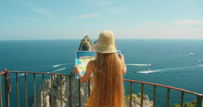 Woman holds map facing iconic Faraglioni cliffs and open Mediterranean sea horizon in Capri Italy