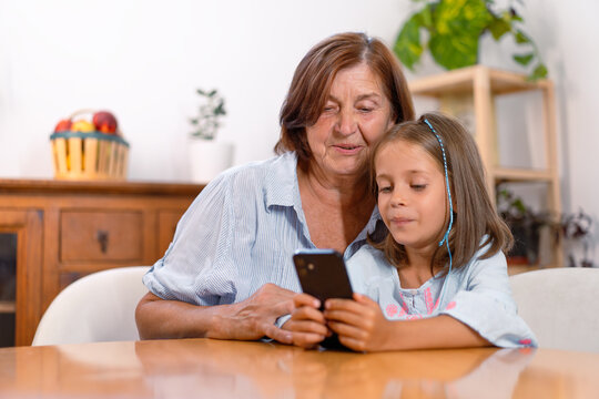 Grandmother and granddaughter using smartphone sitting at table at home, both looking at phone screen, using social media or video calling, enjoying technology together