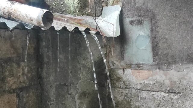 Rainwater pours from a damaged gutter onto an old concrete wall, capturing the raw and gritty atmosphere of heavy rainfall in a worn urban setting