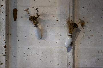 Two white ceramic vases with dried plants hang on a rustic, textured concrete wall, creating a simple, natural, and minimalist decorative display with shadows and earthy tones.