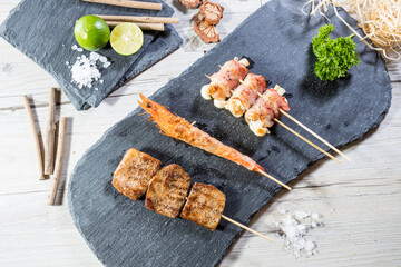 Gourmet skewers of grilled shrimp, bacon-wrapped items, and meat cubes on slate board with lime and coarse salt.