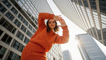 Woman in orange sweater framing face with hands against tall building architecture looking at camera