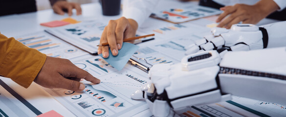 A human hand and a robotic arm collaborate in a contemporary office environment. The scene highlights interaction with charts and data analysis, symbolizing innovation.Noogenesis