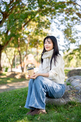 Obraz premium Asian woman in sweater and scarf holding coffee and warming hands as sitting on rock in garden park.