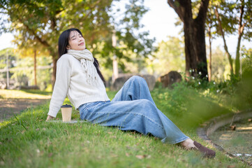 Asian woman in scarf closing eyes leaning back sitting leisurely with coffee on grass in garden park