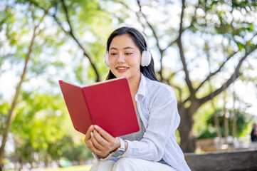Fototapeta premium Asian woman listening to music in headphone and reading a book while sitting on bench in garden park