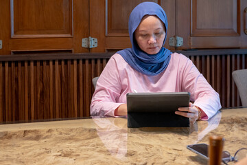 Indonesian muslim woman wearing a blue scarf is sitting at a table with a tablet in front of her. A concept of working in a cafe
