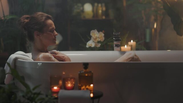 A woman in her 30s finds serene self-care in a luxurious bathtub, surrounded by soft candlelight, lush plants, and steam. With eyes closed and a peaceful expression, she enjoys a tranquil home spa exp