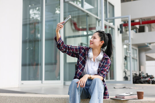 Asian woman student holding smartphone taking picture while sitting on cement floor outside building