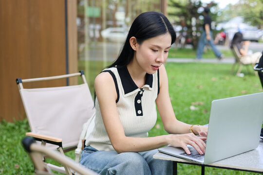 Asian woman student looking and typing on laptop while sitting at table on grass in an outdoors cafe