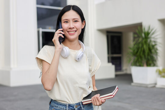 Asian woman student holding book talking on phone walking in front of classroom building or library.
