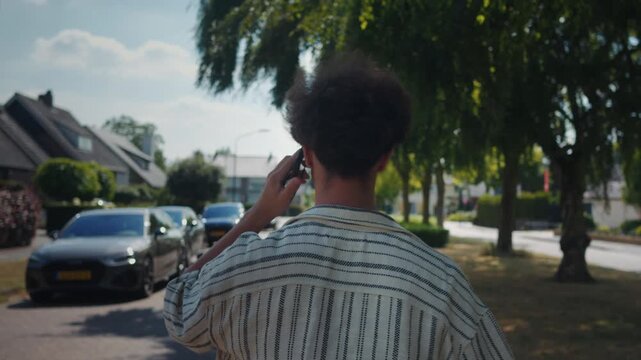 Young man on phone walking past parked cars on suburban street, striped shirt visible from back, animated hand gesture and traffic in background, warm afternoon light evokes everyday