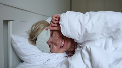 Beautiful positive middle aged woman waking up and peeking out of eye mask at camera with happy smile. Smiling female with cheerful expression woke up in good mood in early morning, lying in comfy bed - Powered by Adobe