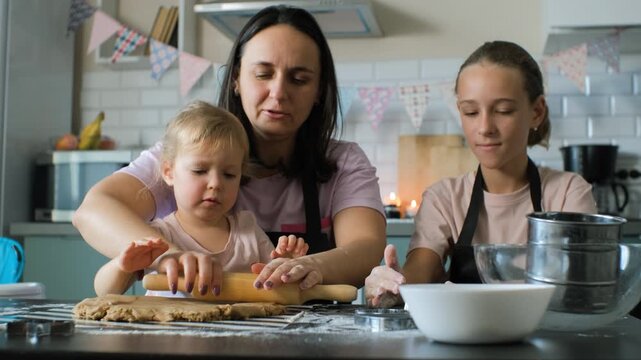 A mother carefully teaches her young daughter how to shape ginger cookie dough at the kitchen table, while the older daughter assists with baking, slow motion