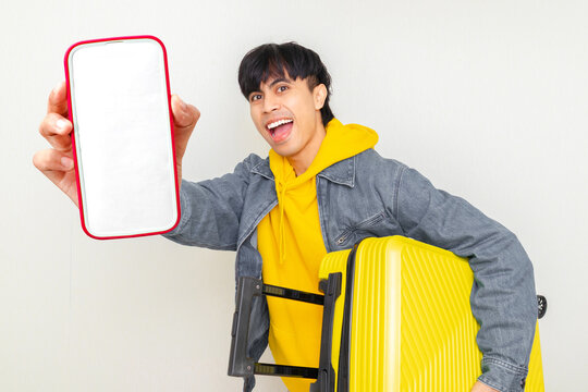 Excited young Asian traveler showing a blank smartphone screen while holding a yellow suitcase, perfect for travel ads, online booking, tourism apps, and vacation promotions.